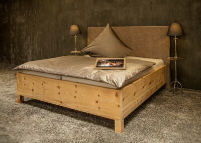 Seitliche Ansicht Massivholz-Bett Rosi. 4cm starker Zirbenholzrahmen auf Blockfüßen mit einem bodentiefen Polsterkopfteil in Wunschfarbe. Rahmenhöhe 35cm, Blockfüße 15cm.